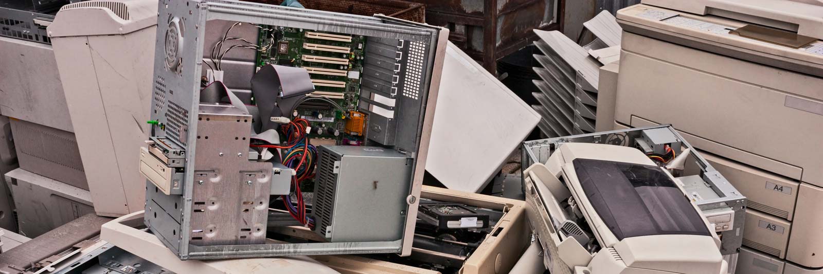Case di un computer smontato con schede e componenti elettronici accanto a stampanti fuori uso, rifiuti RAEE pronti per il trattamento