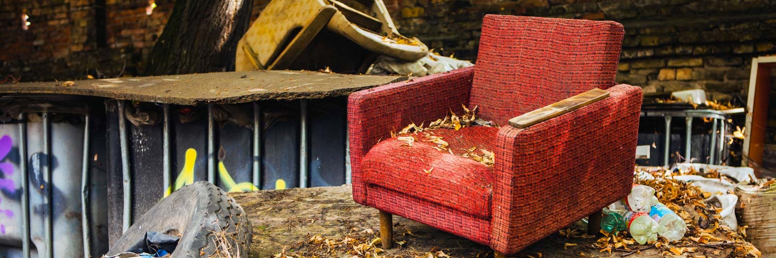 Poltrona rossa vecchia abbandonata all'aperto tra rifiuti e cassonetti, coperta di foglie